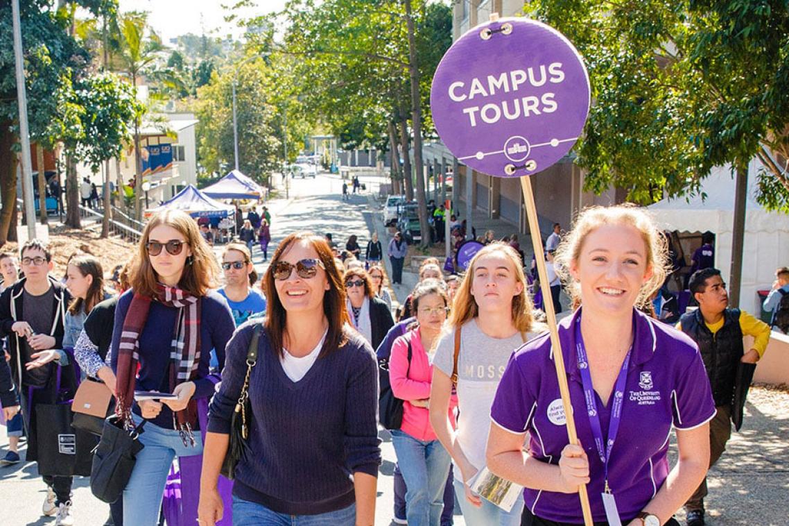 A student in a purple UQ shirt holds a "Campus tours" sign and leads a group of prospective students around St Lucia